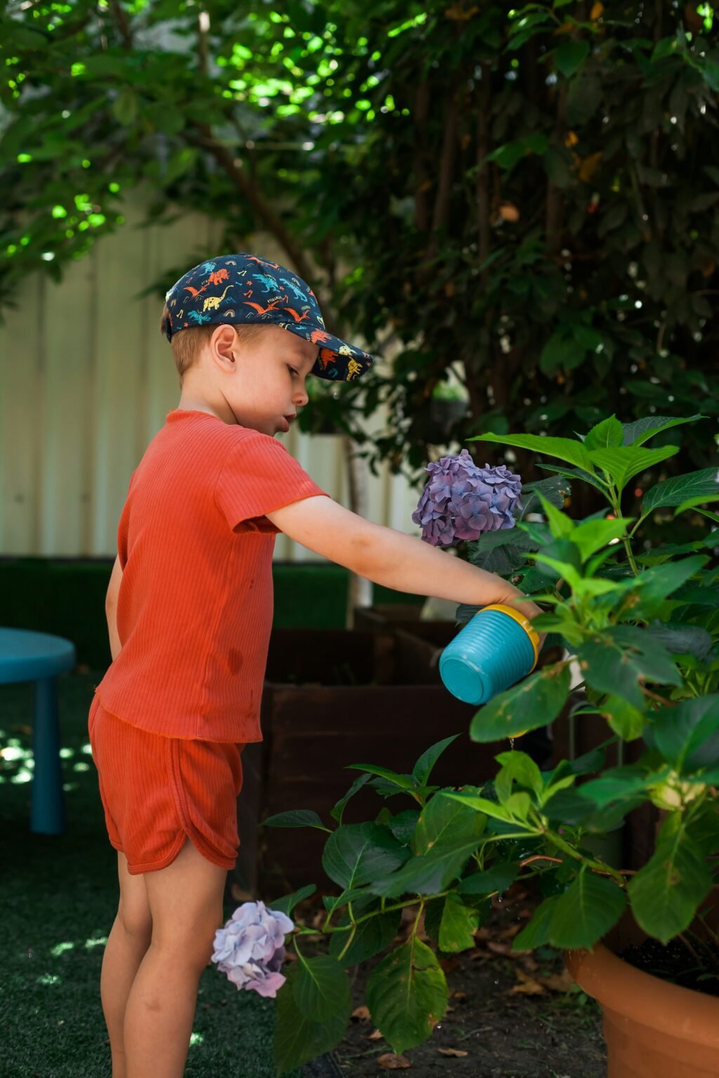 Boy in baseball cap is seen watering plants outdoors.